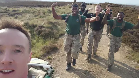 Brendan Hannan Brendan is close to the camera as he is taking the selfie. Five of his friends are in the background walking along a gravel path lined with heather with their thumbs up.