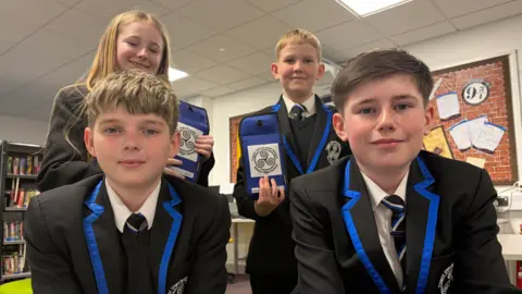 Four pupils - three boys and one girl - in school uniform wearing black blazers with blue edging. Two are sitting by a table and the two standing behind them are holding up phone pouches with the school logo on them.