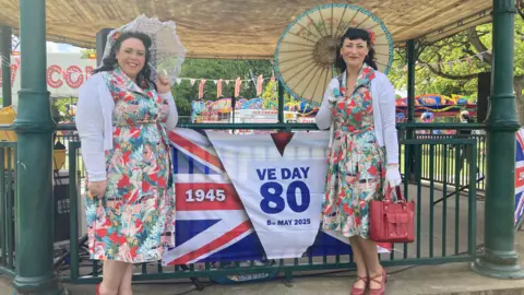 Sally Fairfax/ BBC Two women dressed in vintage floral dresses and white cardigans stood with parasols beside a VE Day 80th anniversary banner. They stand in front of a bandstand at a lively outdoor celebration, with fairground rides and Union Jack bunting in the background.