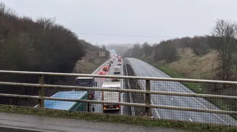 A view of the A47 from a bridge running above it. The left hand carriageway is filled with cars travelling away from the camera in long queues. The right hand carriageway is empty due to the closure.