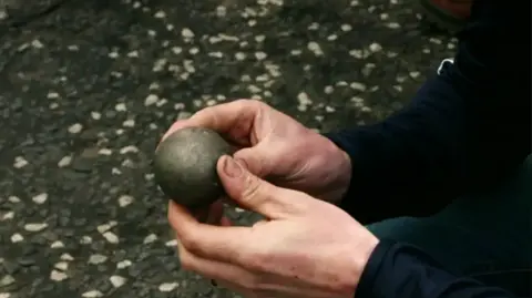 A grey bullet the size of a plum being held between two hands as if the competitor is inspecting it. The competitor's arms and hands are in shot and the stony floor beneath them.  