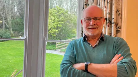 Paul Armogie - a man with a bald head and white short beard. He is wearing glasses and a green jumper with the collar of the shirt underneath popping out