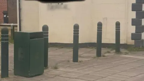 Peter Chaney Graffiti sprayed on a white wall of a housing building. There are words which have been blurred out with a clear blurring. There is a pavement in front of the building and green bollards and a green bin.