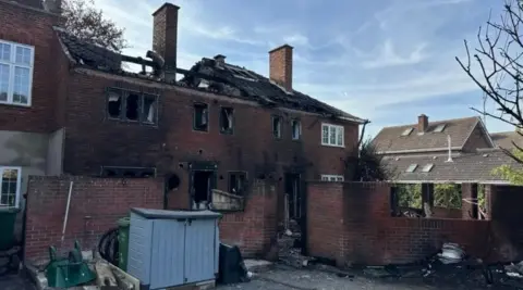 In the centre of the shot is a large, semi-detached home that has been gutted by fire on both floors, with its roof and windows missing. To the left and right are homes unaffected by the blaze. A garden lean-to in a garden at the front is also unaffected.