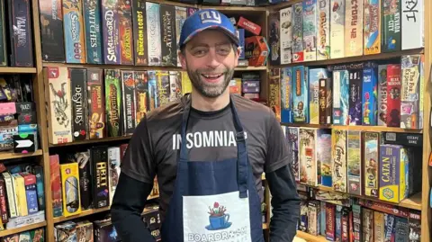 ELLEN KNIGHT/BBC Jon photographed looking into the camera and smiling. He's wearing a long sleeve black top with a brown t-shirt over the top, on which is written 'Insomniac' in white writing. Over that, he's wearing a navy blue apron, which has 'Boardroom' written on it. He's got a bright blue baseball cap on, which has 'NY' embroidered on it in silver letters. He's stood in front of floor-to-ceiling wooden shelves, which have brightly coloured board game boxes stacked on them. 