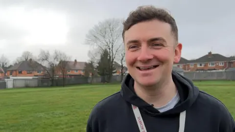 David Moore has short brown hair. He is smiling, he is wearing a blue hoodie and white lanyard with burgundy writing on it. He is standing on a grassy school field and houses can be seen behind him. 