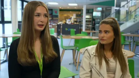Two blonde young women - one in a black cardigan and another in a white jacket - are sitting in a cafe with green chairs positioned at tables.