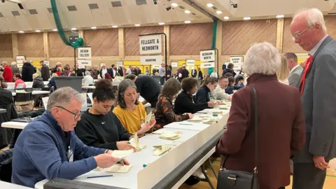 A man wearing a red rosette looks on at the South Tyneside election count. There are rows of people sitting behind desks and counting pieces of paper. There are signs above them with the ward names like: "Fellgate and Hedworth" and "Boldon Colliery".