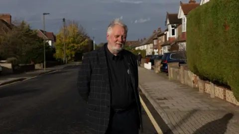 Facundo Arrizabalaga Mr Kent stands in the road with his hands behind his back wearing a black suit with a black shirt. He has grey hair and a short grey beard. There is a green hedge to the right of him.