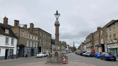 Kate Moser Andon/BBC The broadway in St Ives. View of the Queen Victoria memorial with a large lamp on top, and a street with parked cars and Georgian buildings either side. There are shops including a cycle shop and local cafe.