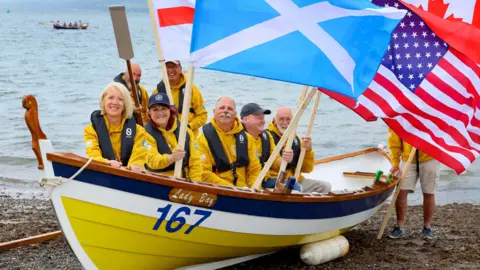 A boat crew dressed in bright yellow wave a range of flags while sitting in a yellow, white and blue boat on the south of Scotland shoreline