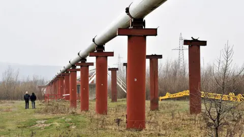 Men stand near the Druzhba crude oil pipeline near Styri, Ukraine in 2009. The pipeline is situated on large orange pillars in a field.