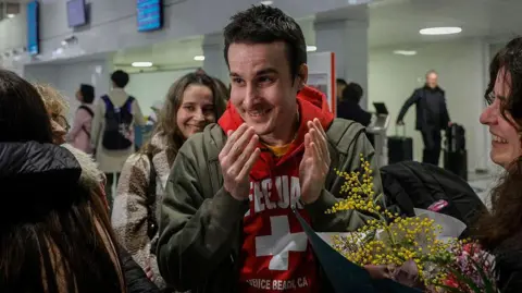 French national Tom Félix is greeted by friends and relatives as he arrives at Roissy-Charles de Gaulle Airport after he was acquitted on eight drug-related charges at the Malaysian High Court. He is smiling while wearing a green jacket and red hoodie which says Lifeguard and has a white plus on it. Several women around him are smiling and one is holding a bunch of flowers.