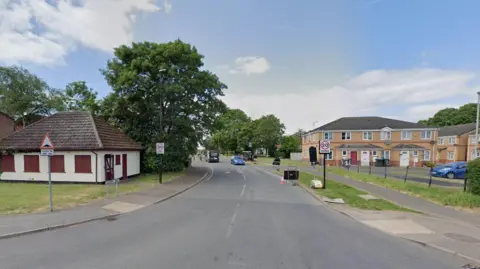 A street view image showing the entrance to a residential street. Buildings are on either side as well as 20mph speed limit signs. 