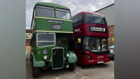 FirstBus Two buses. On the left there is an old green double decker bus, which is showing the number 100 and Sea Mills as the destination. Next to it is a modern red double decker, with citylines written on the front. The destination banner at the top of the bus has a message reading "Lest We Forget, we will remember them"