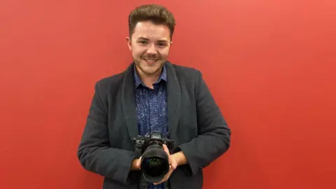 Josh Dury holding a camera and smiling with a red wall behind him