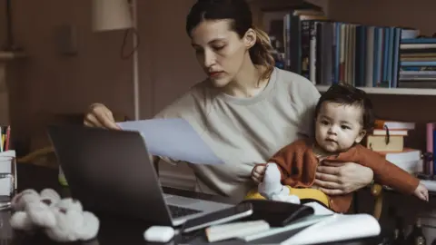 Getty Images Mother holds child as she looks at bills and her computer on a table with a worried expression