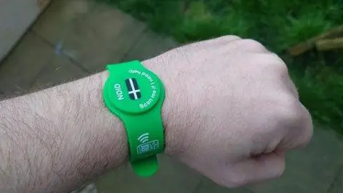 Devon and Cornwall Police A close-up of an arm. The arm has a green wristband on it with a Cornish flag and 'NDID'. Below the hand, in the background, is a patio and the edge of a lawn.