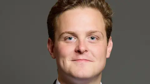 LDRS MP Oliver Ryan, wearing a grey suit and red tie, smiles for the camera in an official photo