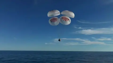 Blue sky with capsule being held up by four parachute-like drogues over the ocean