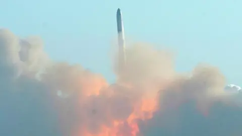 SpaceX's Starship rocket flies vertically into the air against a clear blue sky, leaving behind clouds of smoke and flame. 