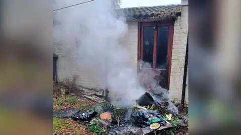 A pile of rubbish on a grassy area in front of a wooden door in a brick wall, with smoke rising from the pile