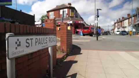 BBC The corner of a  busy high street with a road sign saying St Pauls Road. We can see a red brick wall with square grey paving as the pavement ahead of a road junction.