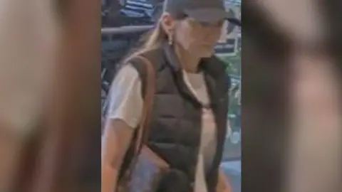 A woman with long hair and wearing a black cap, black gilet and white t-shirt. She is carrying a bag.