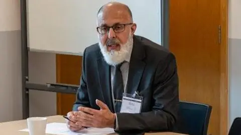 Office of Police, Fire and Crime Commissioner Inam Khan with short dark hair and a white beard and glasses, wearing a dark jacket, light-coloured shirt and dark blue tie, wearing a name badge. He is sitting at the corner of a wooden table with papers and a cup in front of him. There is a wooden door and whiteboard behind him.