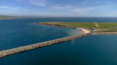 An aerial view of the Churchill Barriers at Scapa Flow. A large expanse of water with a causeway linked to a flat green island.