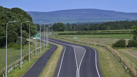 An aerial image of a section of the new Southern Link Road in Carlisle. The single carriage road has a cycle path on one side and is surrounded by green space. It snakes towards the fells in the background.