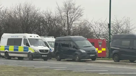 A number of police and black vans on the side of a road, partially on the pavement. There is a red and yellow large van peaking out behind the others. There are leafless trees behind them and the sky is grey.