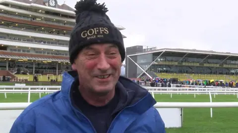 BBC Barry Mason is grinning, looking slightly away from the camera as he gives a TV interview. He is standing on a bit of the racecourse green, with the stands in the background behind him. He wears a black cable knit beanie hat, with the word Goffs in gold embroidery along the front of the hat. He has a high collared blue waterproof coat on with a thick navy blue fleece lining.