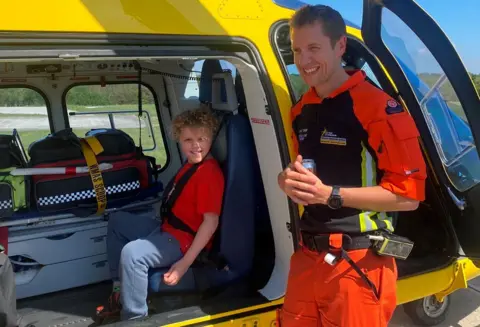 Supplied Boy smiling in red T-shirt with man pictured in Orange suit by the open door of the yellow East Midlands Air Ambulance