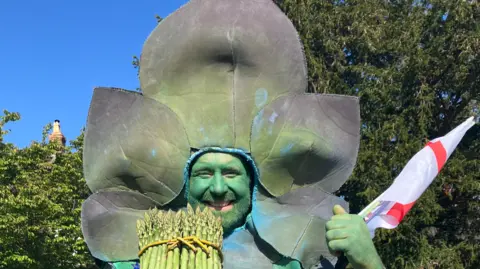 BBC A man with a painted green face is wearing a green asparagus costume and holding asparagus in his right hand. He is smiling and trees are behind him.