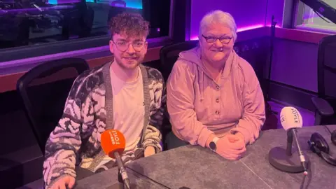 Conor Doherty dalam kaus putih dan cardigan dan Maureen Wilkinson, yang mengenakan hoodie merah muda, digambarkan dalam studio Radio Foyle BBC di Derry. 