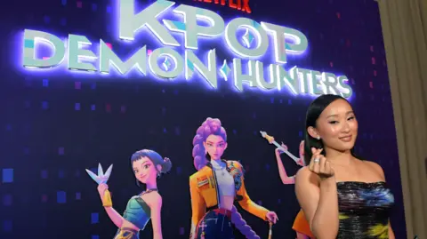 Getty Images SunHee Seo poses in front of a poster for KPop Demon Hunters - the film's logo is lit up with a a purple glow and the three main characters pull poses beneath it on a purple background. Sunhee wears a shoulderless gown and appears to be clicking her fingers as she smiles.