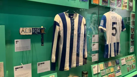 Josh McLaughlin/BBC A retro Brighton & Hove Albion shirt reading 'Dunk' with the number five sits behind a pane of glass. It is surrounded by fanzines, objects and personal collections from the club's history.
