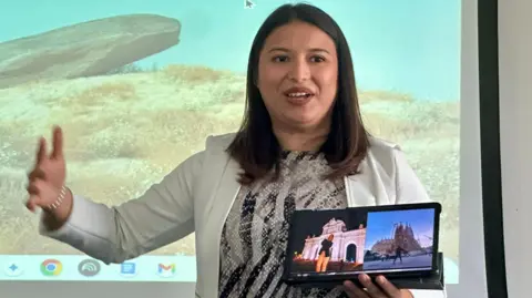 Cad Taylor/BBC A woman is standing in front of a big screen and holding up a tablet with pictures of her home country