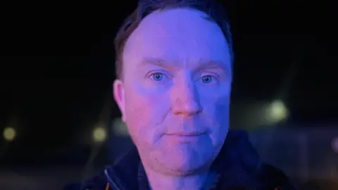 A man stares at the camera in a close up shot. He has very blue eyes and short red hair. 