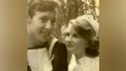 An old black and white photo shows a young man on the left, wearing what appears to be a naval uniform. On the right is a young woman with wavy fair hair, wearing a pale dress and a hat. He is looking at the camera and she is looking up at him.