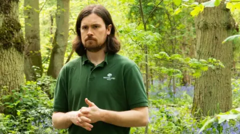 Woodland Trust Jack Taylor, who has shoulder length brown hair and a full beard and moustache, is standing in a bluebell woodland. He is wearing a green polo shirt and has his hands clasped together.