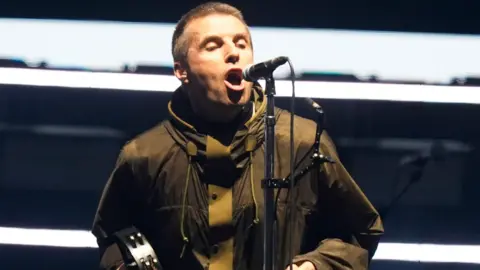 Liam Gallagher with tambourine and khaki jacket in Cardiff, He is singing into a microphone and has short dark hair.