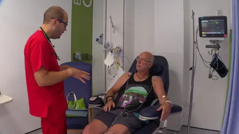 Dr Simon Patten with Andy Mouncey at Royal Devon and Exeter Hospital. Patten is stood wearing red medical scrubs. He is talking with Mouncey, who sitting in a chair with some monitor pads stuck to his shoulders, looking up at Patten.
