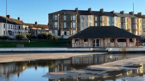 The Boathouse cafe. It is a small white house with black rood and red pillars.  Fairy lights are handing outside and there are tables and chairs.
The empty lake is in front of the building with some puddles from rain water in it. There are residential houses across the street behind the cafe. 