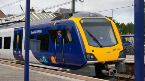 A Northern train which is painted in dark blue and yellow, in a station with the driver looking out of the window.