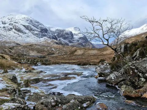 Gordon Pearson A valley stretching between snow-covered mountains. There is a frozen stream in the foreground with brown grasses, rocks and boulders and a tree with bare branches hanging over the water.