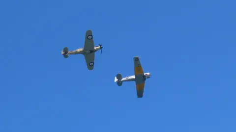 A picture of a Harvard and Hurricane in the sky. There are two planes with the one on the right a grey coloured plane with yellow stripes.