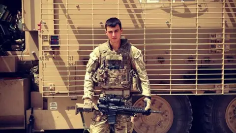 A young Taylor stands in front of an enormous tan-coloured army vehicle. He's wearing camo army gear and holds a large semi-automatic gun. 