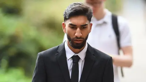 PA Media A young man wearing a black suit and tie and white shirt walks towards the camera. He is looking down with a serious expression. 